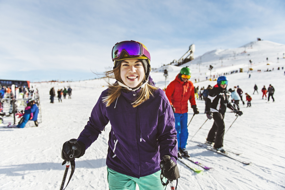 A women standing at the base of the hill, smiling into the camera, with skiers and boarders in the background, before skiing at WinSport in Calgary
